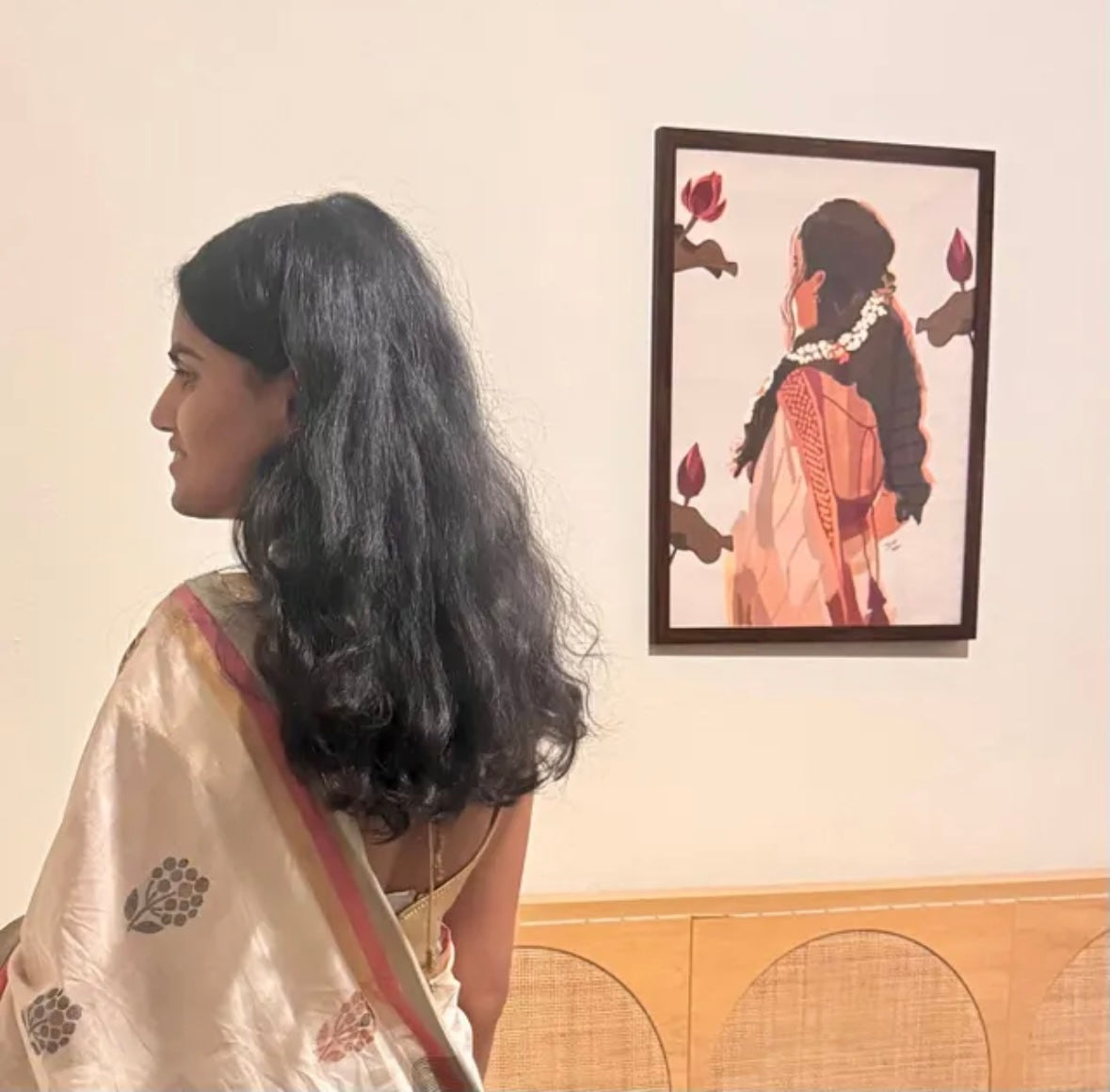 Woman in a white saree looking at a framed artwork of a woman with braided hair and flowers on a beige wall.