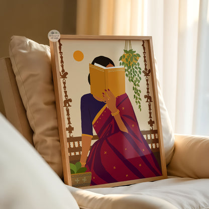A framed art print leaning against a beige sofa in a softly lit room. The print shows a stylized woman in a purple and magenta sari reading a yellow book, with decorative hanging motifs and a cream background.